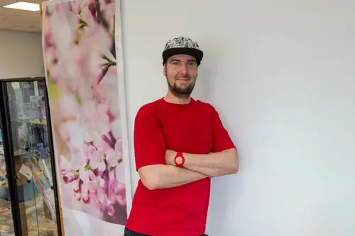 Selbstbewusst lächelnder Mitarbeiter im roten T-Shirt lehnt mit verschränkten Armen an einer weißen Wand.