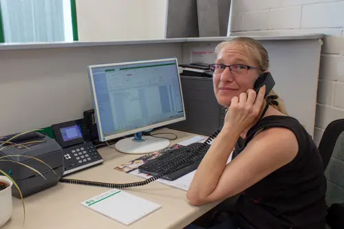 Mitarbeiterin im Kundenservice sitzt an einem Bürotisch, führt ein Telefongespräch und blickt in die Kamera.
